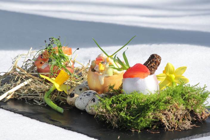 Ein Gourmet-Frühlingsgericht, elegant auf einer Platte präsentiert, mit Wachteleiern, leuchtenden essbaren Blumen und frischen Kräutern auf einem Bett aus Heu und Moos, im Kontrast zum reinweißen Schnee im Hintergrund. - Ihr Landhaus Scherrer aus Hamburg