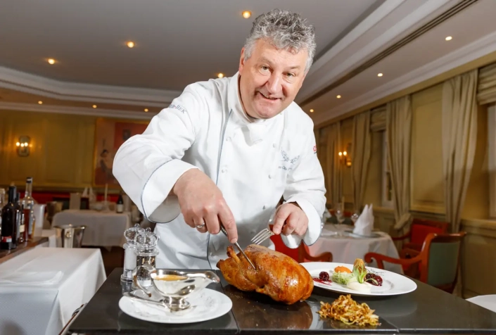 Ein professioneller Koch in weißer Uniform tranchiert im Restaurant Scherrer ein Brathähnchen und ist bereit, in einem eleganten Speisesaal ein Gourmet-Menü zu servieren. - Ihr Landhaus Scherrer aus Hamburg