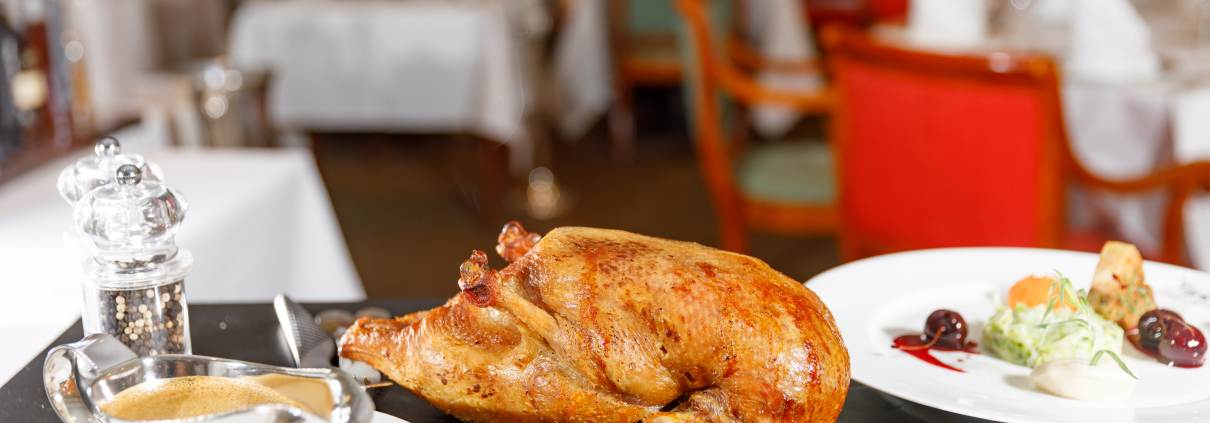 In der Mitte eines eleganten Esstisches steht eine saftige gebratene Ente, dazu eine kleine Sauciere mit Orangen-Pfeffer-Soße, ein Tablett mit winterlichen Gewürzen und eine Beilage aus gemischtem Saisongemüsen. - Ihr Landhaus Scherrer aus Hamburg
