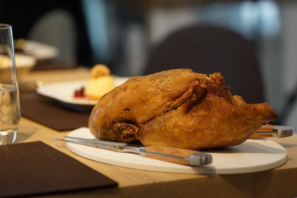 Eine goldbraun gebratene Vierländer Ente auf einem weißen Teller, vor dem unscharfen Hintergrund einer Esstischdekoration im Restaurant Scherrer mit Besteck und Dessertteller. - Ihr Landhaus Scherrer aus Hamburg