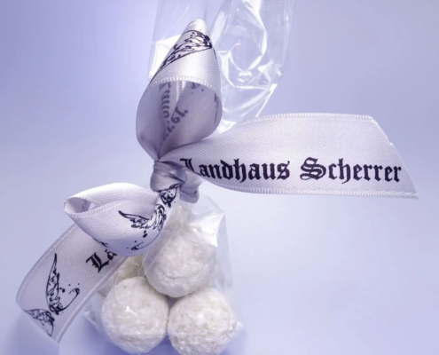 Eine Packung zarter, weiß gepulverter Pralinenselektion-Trüffel, zusammengebunden mit einer eleganten Schleife mit der Aufschrift „Landhaus Scherrer“ und dem sanft bläulichen Hintergrund, der den Eindruck erweckt, es handele sich um eine besondere Köstlichkeit aus einem bestimmten Betrieb. - Ihr Landhaus Scherrer aus Hamburg