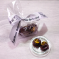 Ein zart mit einer Schleife umwickeltes Paket von Pralinenselektion – handgemacht mit handwerklich hergestellten Pralinen, begleitet von zwei unverpackten Pralinen mit glänzender Oberfläche, die auf einer Glasschale präsentiert werden und ihr verführerisches Wirbeldesign zur Schau stellen. - Ihr Landhaus Scherrer aus Hamburg