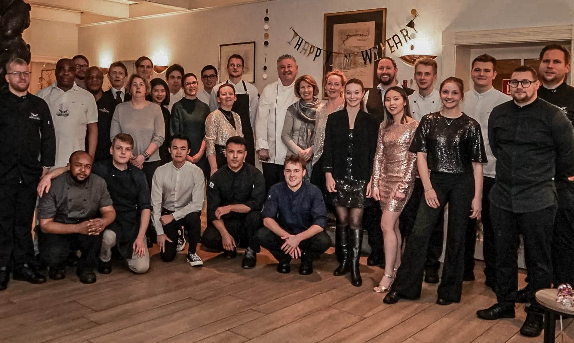 Eine bunt gemischte Gruppe von Menschen, wahrscheinlich das Team vom Landhaus Scherrer, versammelt sich lächelnd und kameradschaftlich unter einem „Frohes neues Jahr!“-Schild für ein Gruppenfoto in Hamburg - Ihr Landhaus Scherrer aus Hamburg