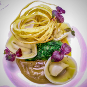 Ein Gourmetgericht mit gefüllten Nudeln, einer leuchtend grünen Soße, knusprig frittierten Zwiebelringen und garniert mit zarten violetten Blüten, wunderschön präsentiert auf einem weißen Teller mit violettem Akzent. - Ihr Landhaus Scherrer aus Hamburg Ein Gourmetgericht mit gefüllten Nudeln, einer leuchtend grünen Soße, knusprig frittierten Zwiebelringen und garniert mit zarten violetten Blüten, wunderschön präsentiert auf einem weißen Teller mit violettem Akzent. - Ihr Landhaus Scherrer aus Hamburg