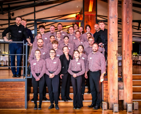 Ein gut gekleidetes Mitarbeiterteam in passenden karierten Uniformen versammelt sich für ein Gruppenfoto in einem warmen, mit Holzakzenten versehenen Veranstaltungsort und demonstriert so Zusammenhalt und die Bereitschaft, außergewöhnlichen Service zu bieten. - Ihr Landhaus Scherrer aus Hamburg