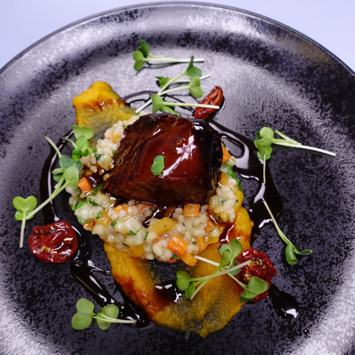 Gourmet-Gericht mit glänzend glasiertem Protein auf einem Bett aus cremigem Risotto, garniert mit Microgreens, sonnengetrockneten Tomaten und einem farbenfrohen Kürbispüree, serviert auf einem dunklen Keramikteller. - Ihr Landhaus Scherrer aus Hamburg