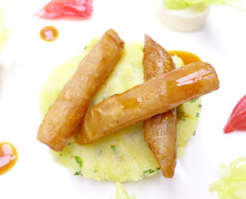 Goldbraune karamellisierte Bananen auf einem Bett aus leuchtend grünem, mit Kräutern angereichertem Kartoffelpüree, garniert mit zarten grünen Blättern und kunstvollen Tupfen aus roter und orangefarbener Sauce, präsentieren eine Fusion aus süßen und herzhaften Aromen. - Ihr Landhaus Scherrer aus Hamburg