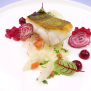 In der Pfanne gebratenes Fischfilet mit goldbrauner, knuspriger Haut, begleitet von einer Mischung aus Birnenwürfeln, Kirschreduktionen und leuchtend roten Scheiben von violettem Rettich, kunstvoll präsentiert auf einem weißen Teller. - Ihr Landhaus Scherrer aus Hamburg