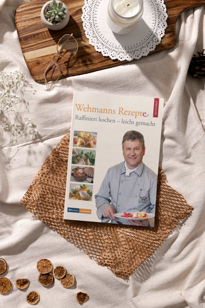 Ein Kochbuch mit dem Titel „Wehmanns Rezepte“ wird auf einem strukturierten Stoff neben kulinarischen Zutaten und Küchengeschirr präsentiert und suggeriert eine Szene der gastronomischen Zubereitung und raffinierten Küche rund um traditionelle deutsche Gerichte - Ihr Landhaus Scherrer aus Hamburg