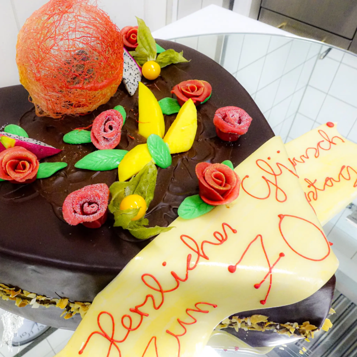 Ein wunderschön dekorierter Schokoladenkuchen, geschmückt mit farbenfrohen essbaren Blumen und einem zarten Zuckernetz, abgerundet durch ein festliches gelbes Banner mit einer handgespritzten Inschrift zur Feier eines 70. Geburtstags im Landhaus Scherrer. - Ihr Landhaus Scherrer aus Hamburg