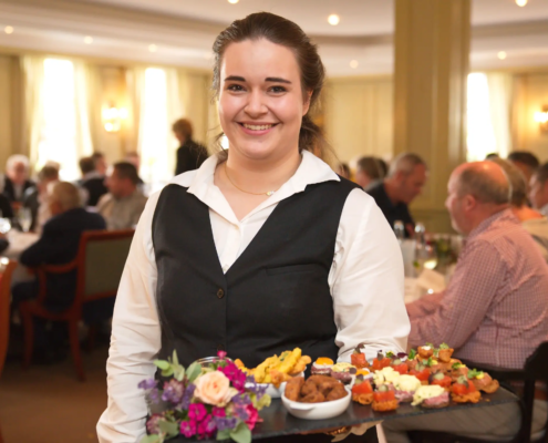 In einem geschäftigen Restaurant präsentiert ein lächelnder Kellner eine Platte mit Vorspeisen und heißt die Gäste, die im Hintergrund ihr Essen genießen, herzlich willkommen. - Ihr Landhaus Scherrer aus Hamburg