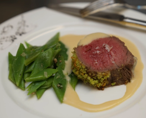 Ein saftiges, medium gebratenes Rinderfilet mit einer Kräuterkruste, dazu knackig-zarte grüne Bohnen, beträufelt mit einer seidigen, herzhaften Sauce auf einem weißen Teller, garniert mit Flockensalz. - Ihr Landhaus Scherrer aus Hamburg