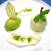 Ein weißer Teller mit einem raffinierten Dessertarrangement aus dem renommierten Restaurant von Heinz O. Wehmann. Links eine Kugel grünes Matcha-Eis, geschmückt mit einer zarten, goldenen Zuckerdekoration. Rechts ein weißes, rundes Gebäck, darauf ein Minzzweig. Darauf sind kunstvoll Sahnesaucen-Spiralen mit grünen Streuseln verteilt. - Ihr Landhaus Scherrer aus Hamburg