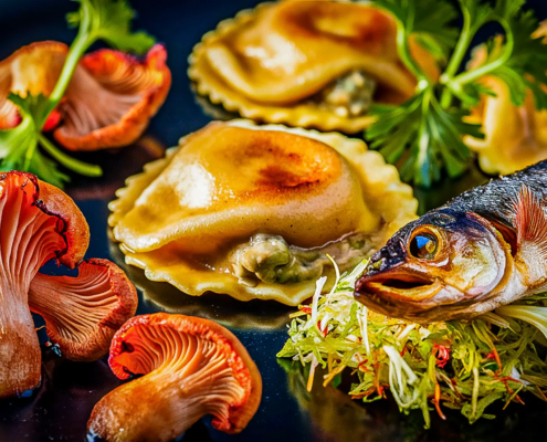 Ein Gourmetgericht im Restaurant Hamburg, zubereitet von Chefkoch Heinz O. Wehmann, mit gefüllten Nudeln, wahrscheinlich Ravioli, angerichtet auf einem schwarzen Teller mit frischen Korianderzweigen. Überall sind farbenfrohe Pfifferlinge verstreut und ein ganzer gekochter Fisch ruht auf einem Bett aus fein geschnittenem Grünzeug. - Ihr Landhaus Scherrer aus Hamburg
