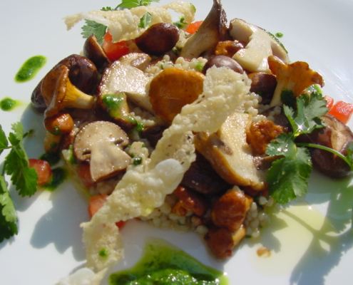 Herbstlich angerichtetes Graupenrisotto mit einer Auswahl gebratener Pilze, darunter Steinpilze, Pfifferlinge und Champignons. Das Gericht ist mit knusprigen Parmesan-Chips, frischer Petersilie, kleinen Tomatenwürfeln und grünem Petersilienöl garniert und auf einem weißen Teller angerichtet.